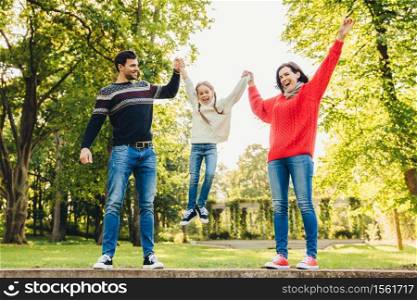 Funny playful child rocks in parent`s hands, being overjoyed. Father and mother play with their little daughter outdoors. Concept of happy family. Parents hold child`s hands. Parenthood and people