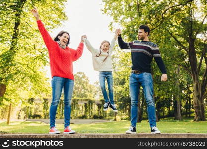 Funny playful child rocks in parent`s hands, being overjoyed. Father and mother play with their little daughter outdoors. Concept of happy family. Parents hold child`s hands. Parenthood and people