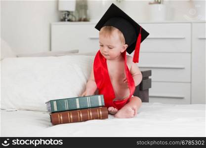 Funny portrait of 10 months baby boy in graduation cap looking at big books