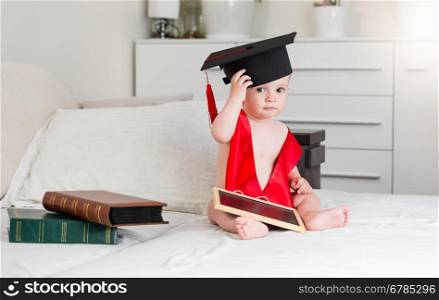 Funny portrait of 10 months old baby boy with books wearing graduation cap