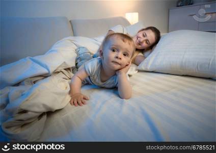 Funny portrait of adorable baby boy lying on bed next to his mother trying to sleep