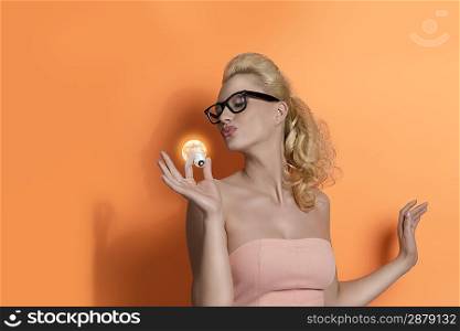 funny portrait of blonde girl with pink dress and black glass. She kissing light bulb on orange background