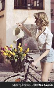 funny portrait of sexy blonde woman with denim shorts watering coloured tulips in the bicycle?s basket