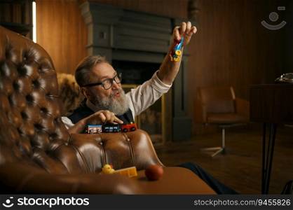 Funny senior businessman playing toys enjoying fun time after hard working day in home office. Elderly male sitting on floor nearby armchair. Funny senior businessman playing toys enjoying fun time after hard working day