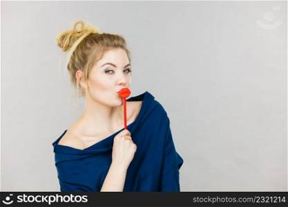 Funny woman holding big red lips on stick. Blonde young female ready for party, on grey background. Humor costume, smart and carnival concept.. Funny woman holding big red lips on stick