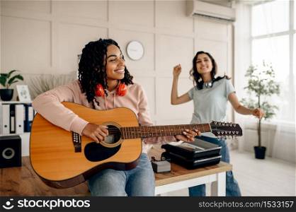 Funny women play the guitar and listen to music at home. Pretty girlfriends in earphones relax in the room, sound lovers resting on couch, female friends leisures together. Women play the guitar and listen to music