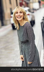 Funny young blonde woman standing on urban background. Smiling blonde girl wearing dress dancing outdoors.