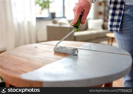 furniture renovation, diy and home improvement concept - close up of woman in gloves with paint roller painting old wooden table in grey color. woman painting old wooden table in grey color