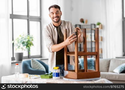 furniture renovation, diy and home improvement concept - happy smiling man sanding old wooden table or chair with sponge. man sanding old round wooden table with sponge
