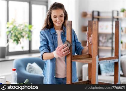 furniture renovation, diy and home improvement concept - happy smiling woman sanding old wooden table or chair with sponge. woman sanding old round wooden table with sponge