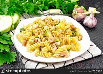 Fusilli pasta with chicken breast, zucchini, cream and pine nuts in a plate on napkin, garlic, fork and parsley on a dark wooden board background