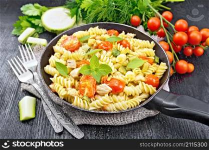 Fusilli with chicken, zucchini and tomatoes in a frying pan on burlap, forks, basil and parsley on wooden board background