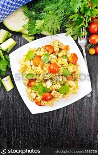 Fusilli with chicken, zucchini and tomatoes in a plate, napkin, fork, basil and parsley on wooden board background from above