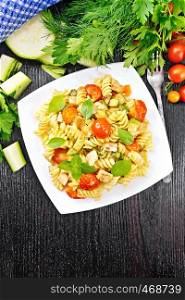 Fusilli with chicken, zucchini and tomatoes in a plate, napkin, fork, basil and parsley on wooden board background from above
