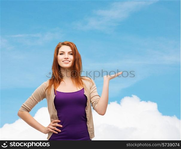 future, dream, advertising and people concept - smiling teenage girl in casual clothes holding something on her palm over blue sky and cloud background