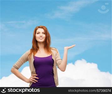 future, dream, advertising and people concept - smiling teenage girl in casual clothes holding something on her palm over blue sky and cloud background