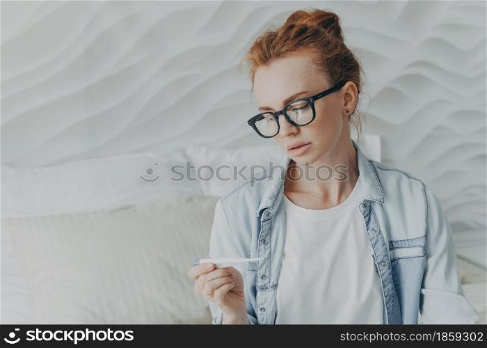 Future mother with natural red hair looks attentively at positive pregnancy test result finally gets pregnant checks ovulation test wears spectacles denim shirt poses against cozy domestic interior. Future mother with natural red hair looks attentively at positive pregnancy test result