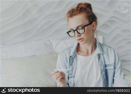Future mother with natural red hair looks attentively at positive pregnancy test result finally gets pregnant checks ovulation test wears spectacles denim shirt poses against cozy domestic interior. Future mother with natural red hair looks attentively at positive pregnancy test result
