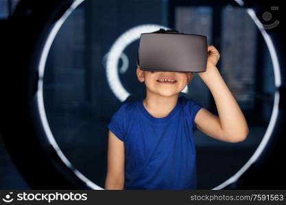 future technology and people concept - happy smiling boy vr glasses or virtual reality headset over white illumination in dark room. boy in vr glasses over illumination in dark room