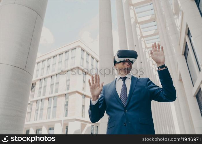 Futuristic image of happy caucasian businessman using virtual reality 3d goggles while standing outdoors, managing project in cyberspace, testing VR headset with blurred buildings in background. Futuristic image of happy businessman using virtual reality 3d goggles while standing outdoors