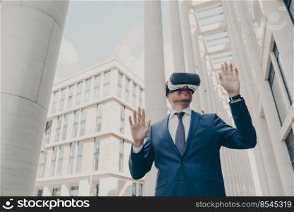Futuristic image of happy caucasian businessman using virtual reality 3d goggles while standing outdoors, managing project in cyberspace, testing VR headset with blurred buildings in background. Futuristic image of happy businessman using virtual reality 3d goggles while standing outdoors