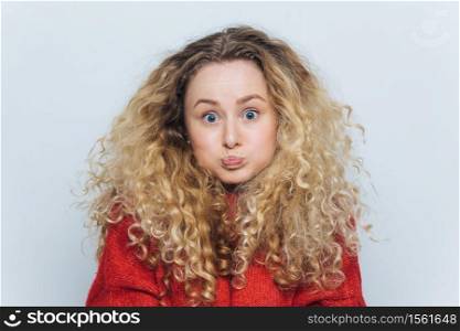 Fuuny blonde female with curly hair, blows cheeks and stares at camera, reacts actively on unexpected news, isolated over white background. Emotional young woman poses indoor. Body language concept