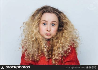 Fuuny blonde female with curly hair, blows cheeks and stares at camera, reacts actively on unexpected news, isolated over white background. Emotional young woman poses indoor. Body language concept