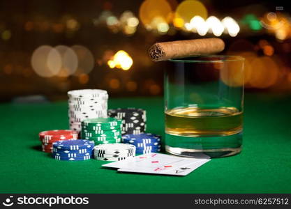 gambling, fortune and entertainment concept - close up of casino chips, whisky glass, playing cards and cigar on green table surface over holidays night lights background