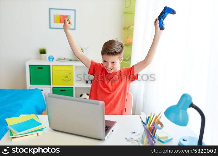 gaming, technology and people concept - smiling boy with gamepad and laptop computer playing video game and celebrating victory at home. boy with gamepad playing video game on laptop