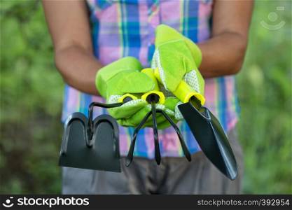 Garden tools in the hands of a woman farmer. Women's gloved hands hold a shovel, chopper and rake. Green natural background. Garden tools in the hands of woman farmer