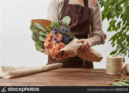Gardener woman with bouquet for a holiday from living coral roses and branches of green leaf on a wooden table on a light background, place for text. Mother's Day holiday.. Creating of beautiful flowers bouquet from living coral color roses with green leaves in a paper in a woman's hands with tattoo on a gray background.