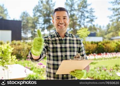 gardening and people concept - happy smiling middle-aged man in apron with clipboard showing thumbs up at summer garden. man with clipboard showing thumbs up at garden