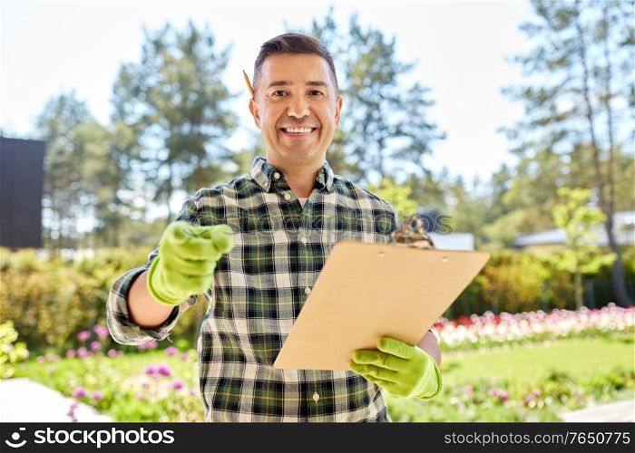 gardening and people concept - happy smiling middle-aged man in apron with clipboard pointing to camera at summer garden. man with clipboard pointing to camera at garden