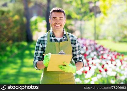 gardening and people concept - happy smiling middle-aged man in apron writing to clipboard at summer garden. happy man with clipboard at summer garden