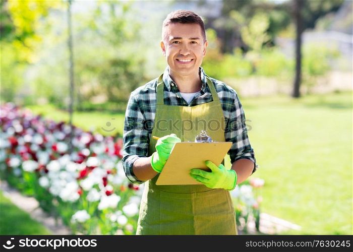gardening and people concept - happy smiling middle-aged man in apron writing to clipboard at summer garden. happy man with clipboard at summer garden