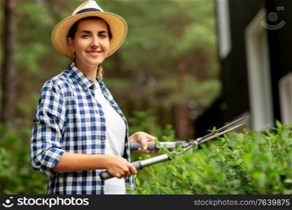 gardening and people concept - happy smiling young woman in straw hat with pruner or pruning shears cutting branches at summer garden. woman with pruner cutting branches at garden