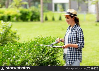 gardening and people concept - happy smiling young woman in straw hat with pruner or pruning shears cutting branches at summer garden. woman with pruner cutting branches at garden