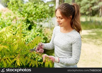 gardening and people concept - happy young woman or gardener with pruner taking care of bushes at summer garden. woman with pruner cutting bushes at summer garden