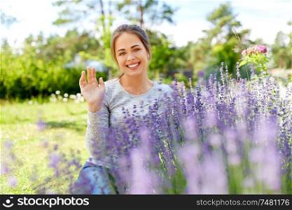 gardening and people concept - happy young woman sitting near lavender flowers on summer garden bed and waving hand. young woman and lavender flowers at summer garden