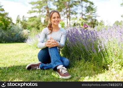 gardening and people concept - happy young woman sitting on grass near lavender flowers on summer garden bed. young woman and lavender flowers at summer garden