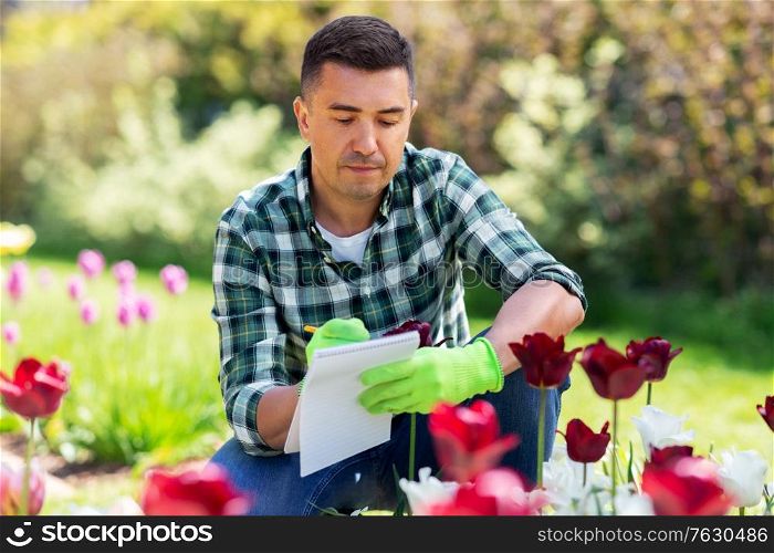 gardening and people concept - middle-aged man writing to notebook and taking care of flowers at summer garden. man with notebook and flowers at summer garden