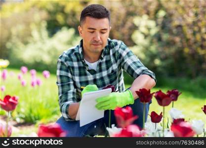gardening and people concept - middle-aged man writing to notebook and taking care of flowers at summer garden. man with notebook and flowers at summer garden