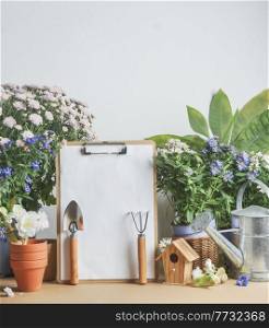 Gardening background with empty to do list, gardening equipment and potted flowers on table at white wall background. Front view with copy space.