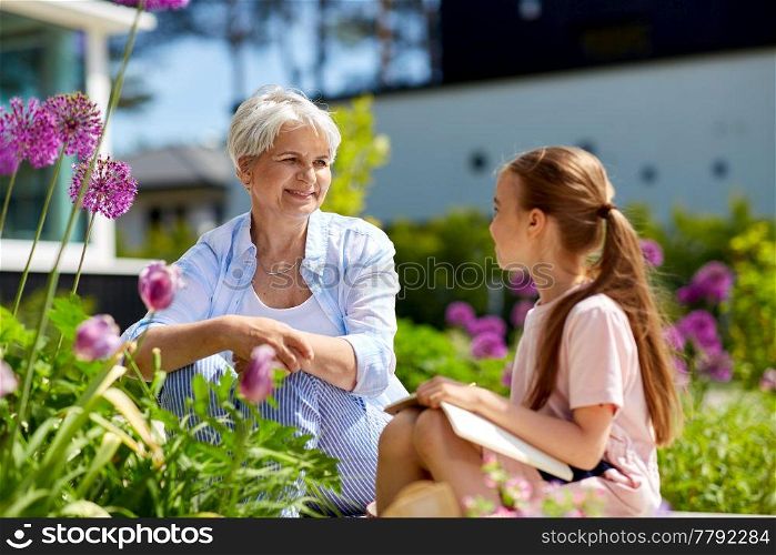 gardening, family and people concept - happy grandmother and granddaughter with notebook and flowers at summer garden. grandmother and granddaughter at summer garden