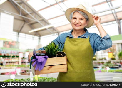 gardening, farming and old people concept - portrait of smiling senior woman in green apron and straw hat holding wooden box with garden tools over greenhouse at garden store on background. senior woman with tools in box at garden store