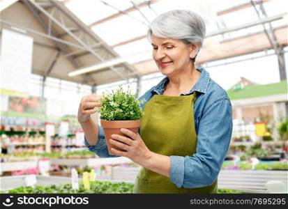 gardening, farming and old people concept - portrait of smiling senior woman in green apron with flower in pot over greenhouse at garden store on background. smiling senior woman with flower at garden store