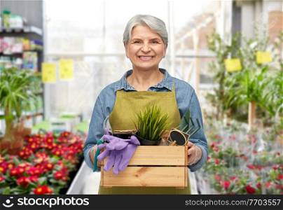 gardening, farming and old people concept - portrait of smiling senior woman in green apron holding wooden box with garden tools over greenhouse with flowers background. smiling senior woman with garden tools in box