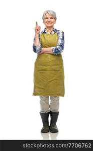 gardening, farming and old people concept - portrait of smiling senior woman in green garden apron and rubber boots pointing finger up over white background. happy old woman in garden apron pointing finger up