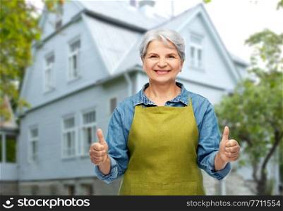 gardening, farming and old people concept - portrait of smiling senior woman in green garden apron showing thumbs up over house background. senior woman in garden apron showing thumbs up