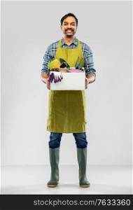 gardening, farming and people concept - happy smiling indian male gardener or farmer in apron and rubber boots with box of garden tools over grey background. indian gardener or farmer with box of garden tools